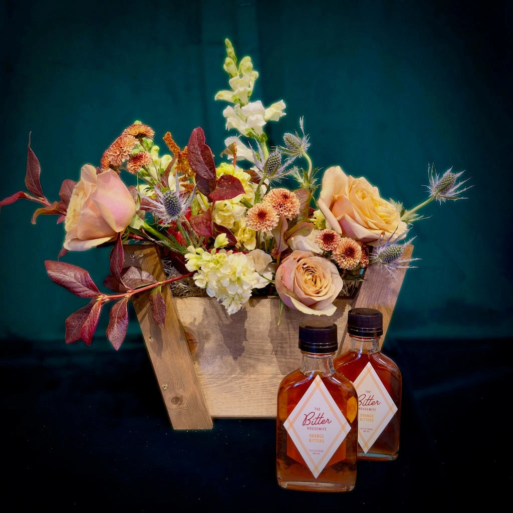 A custom curated gift basket including a seasonally inspired floral design accompanied by two "The Bitter Housewife" bottles of bitters. Designed in a handmade wooden gift basket by Seattle Florist Campanula Design Studio