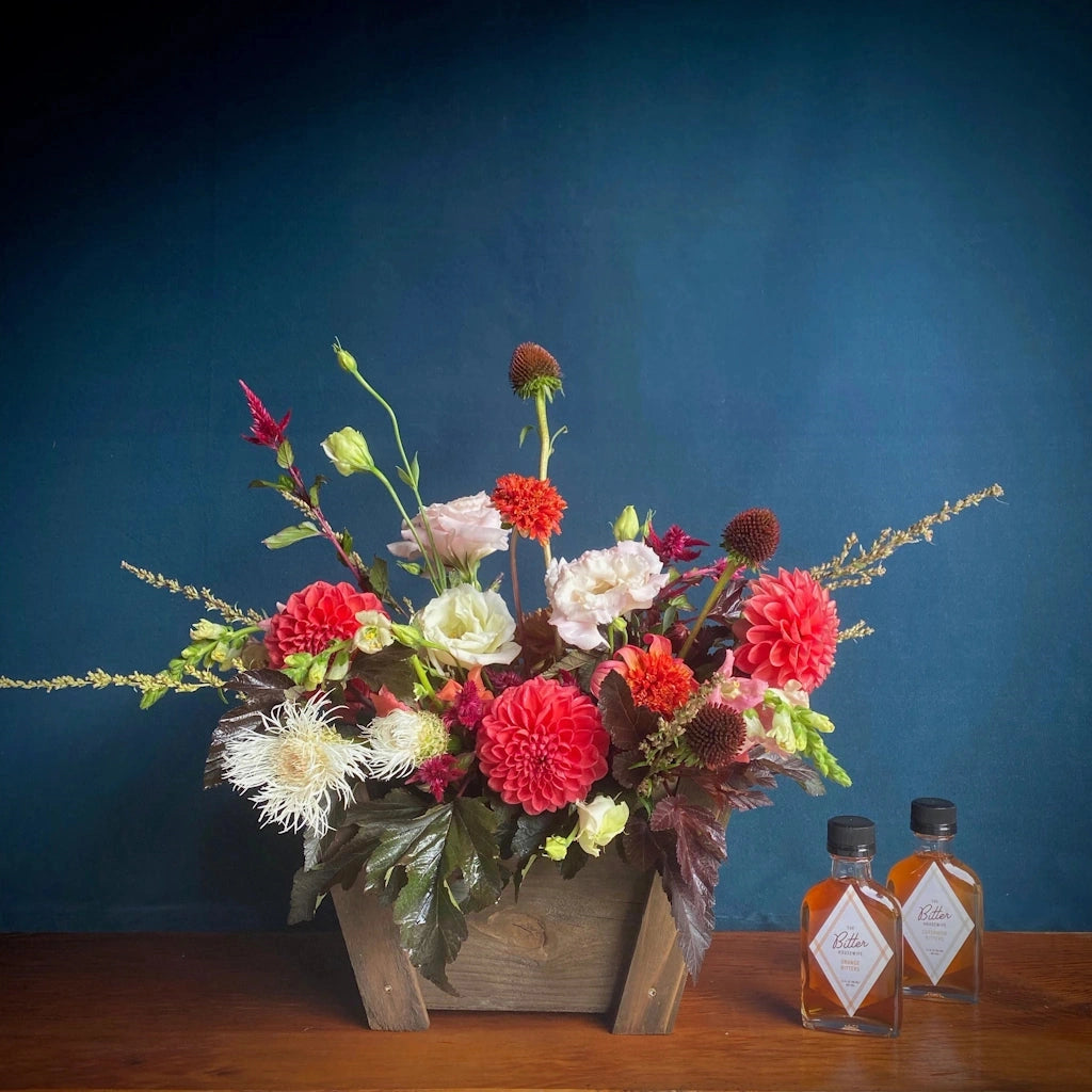 A custom curated gift basket including a seasonally inspired floral design accompanied by two "The Bitter Housewife" bottles of bitters. Designed in a handmade wooden gift basket by Seattle Florist Campanula Design Studio